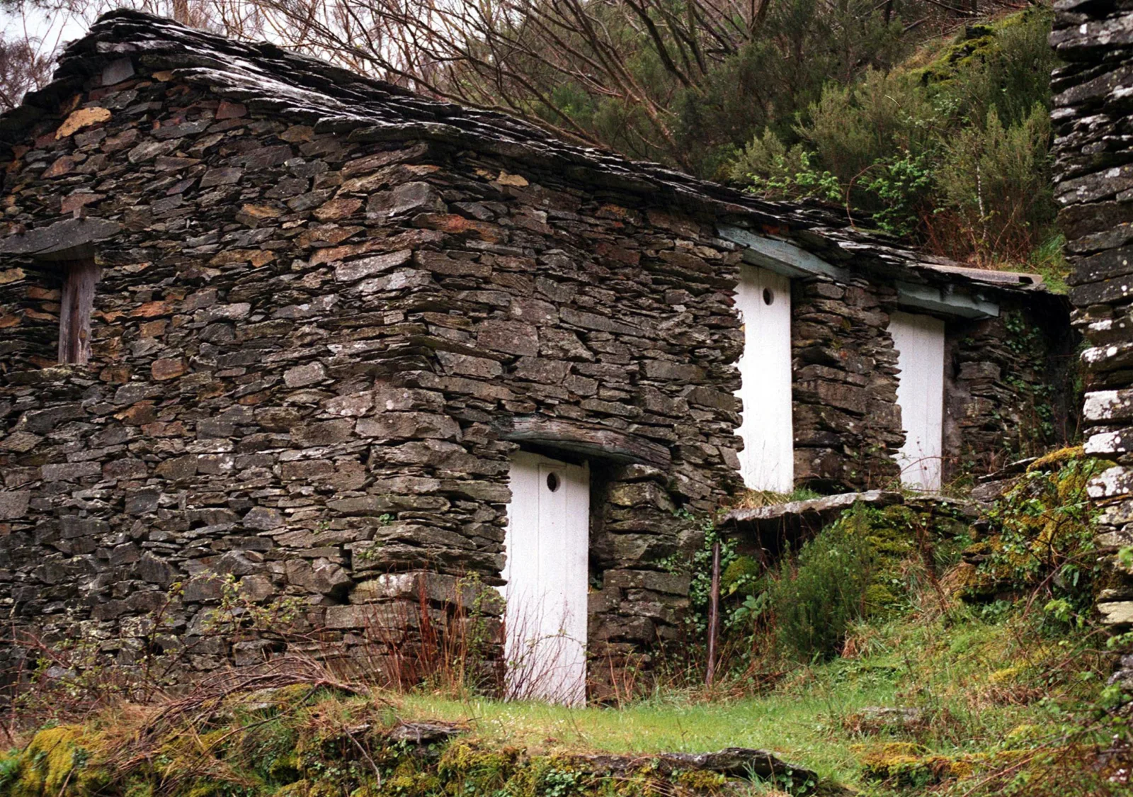 Construções rústicas de xisto com portas brancas, rodeadas de vegetação, na aldeia histórica de Piódão, em Portugal.