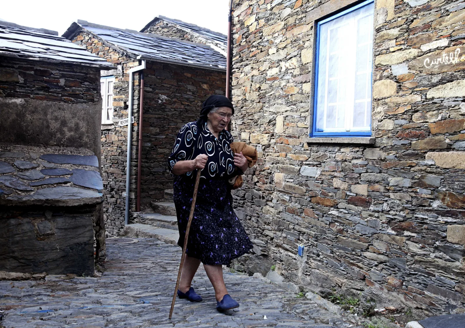 Mulher idosa caminha por rua de pedra na aldeia histórica de Piódão, em Portugal, segurando um cajado e um xaile.